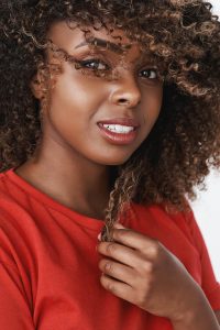 Close-up shot of romantic and sensual gorgeous dark-skinned girl with pure clean skin smiling tender touching curl of hair and waving haircut gazing soft and caring at camera over white background.