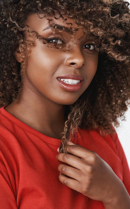 Close-up shot of romantic and sensual gorgeous dark-skinned girl with pure clean skin smiling tender touching curl of hair and waving haircut gazing soft and caring at camera over white background.